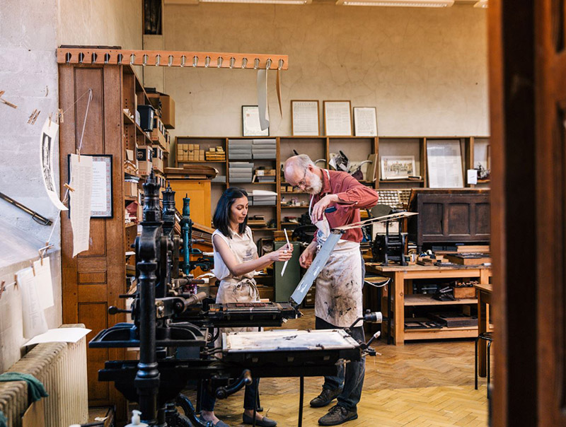Two people look at a print in front of an old printing press