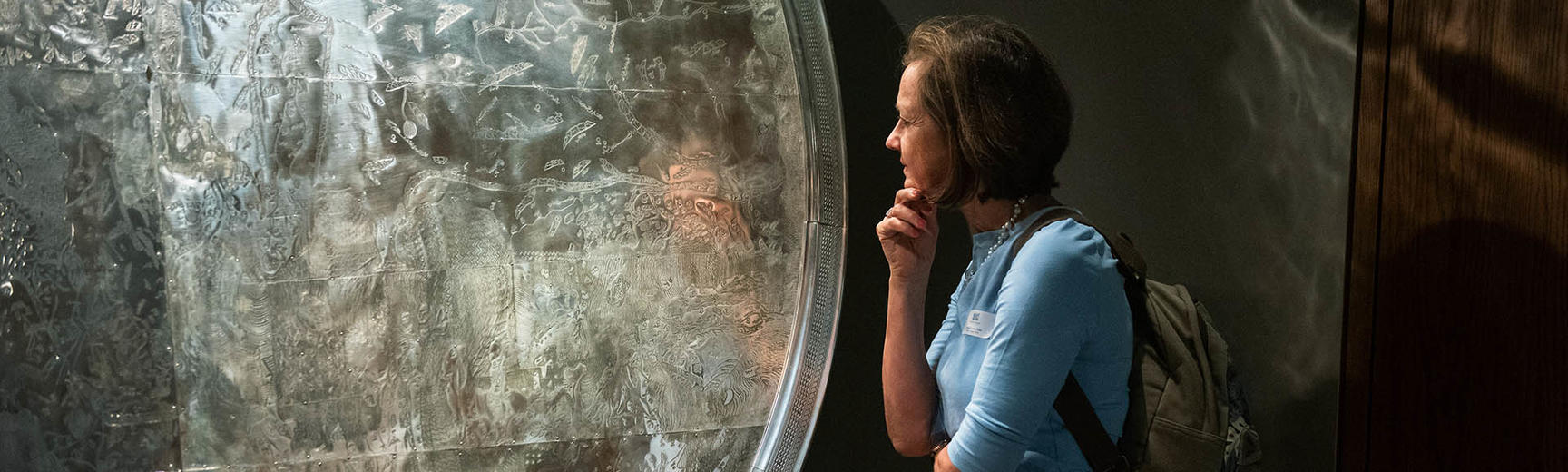 A woman wearing a blue jumper and a backpack stands in front of a large map