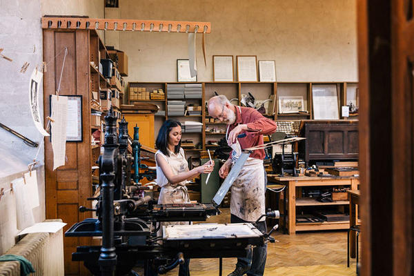 Two people look at a print in front of an old printing press