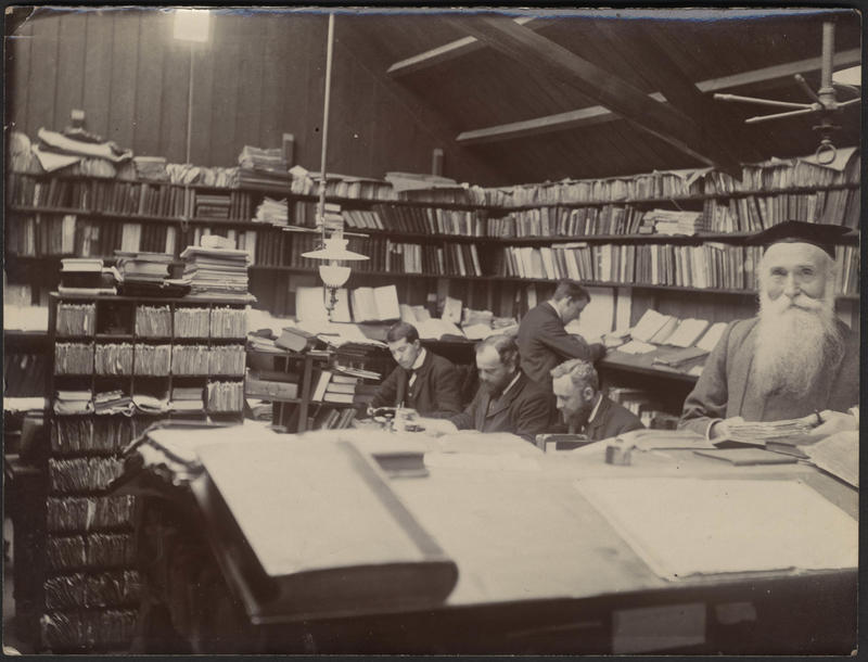 A black and white photograph of a man with a long white beard holding a book, in the background four men work at desks surrounded by books